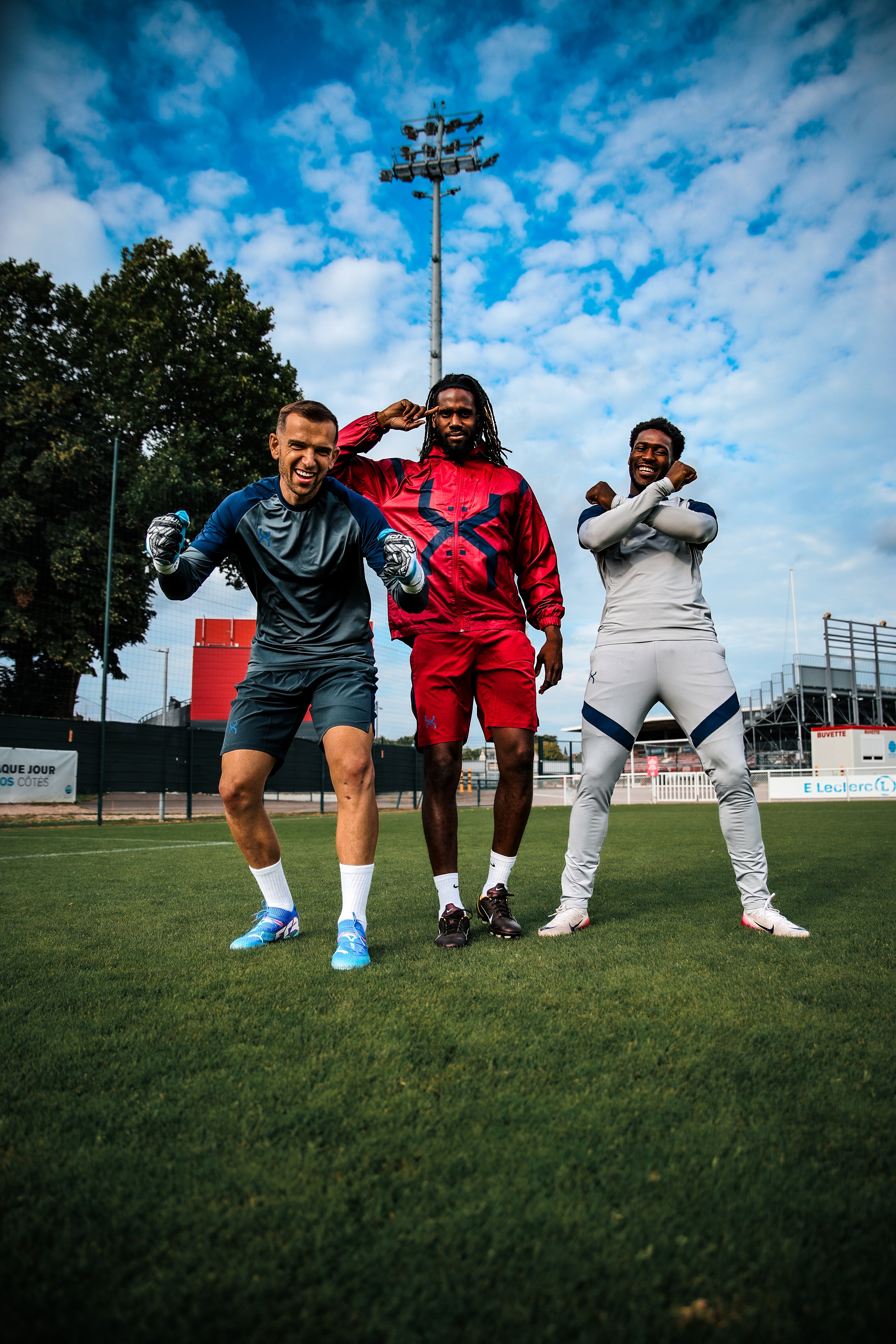 Three individuals posing on a soccer field with a blue sky and clouds in the background.